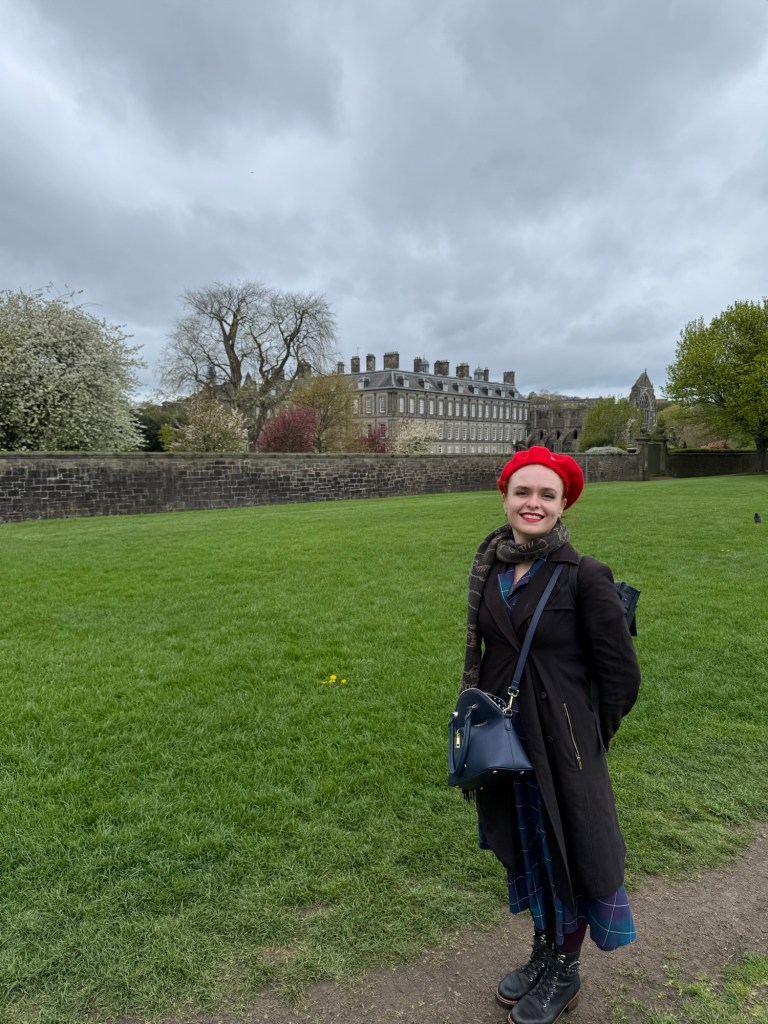 A woman wearing a red beret poses in front of a green lawn with the Edinburgh palace in the background