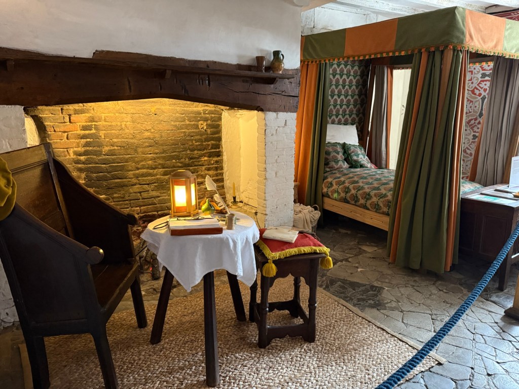A table and chair sit next to a hearth with a four-poster bed to the side