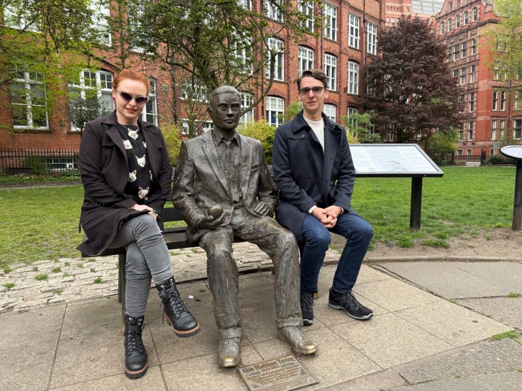 Two people sit side by side with a bronze statue of Alan Turing in the middle