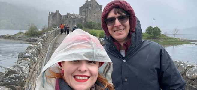 Two people in raincoats pose in front of a castle